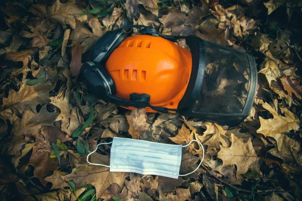 フォレスト内の乾燥した葉の背景にある抗ウイルスマスクの隣に働くヘルメット — ストック写真