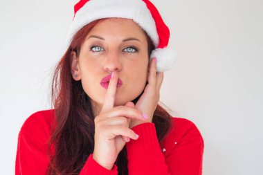 Kırmızı bir süveter ve Noel Baba şapkası giyen dudaklarına parmağını dayayan bir kadın.