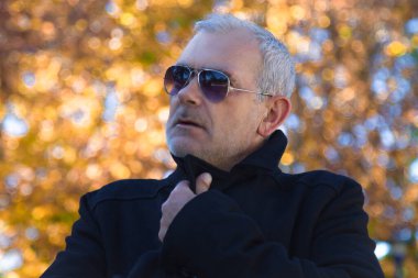 Sonbahar parkında siyah ceket ve güneş gözlüğü takan yakışıklı bir adam.