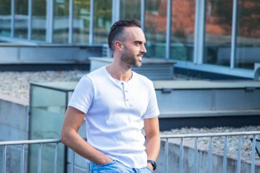 Gömlek ve kot giymiş genç bir adam, elleri ceplerinde sokakta dikiliyor, rahat ve kendinden emin bir görünüm sergiliyor..