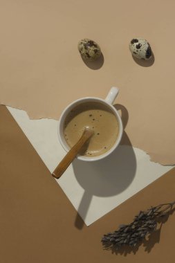 Paskalya pastası ve bıldırcın yumurtalarıyla sert bir gölgeyle kumlu bej arka planda bir fincan latte. Paskalya kartı minimalizmi. Moda izometrik bej arka plan. Yaratıcı kartpostal