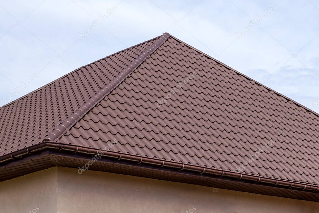 Roof structure covered with brown metal tiles on a bright sunny day. Modern housing construction. Types of hipped roof and valley roofs. House exterior.