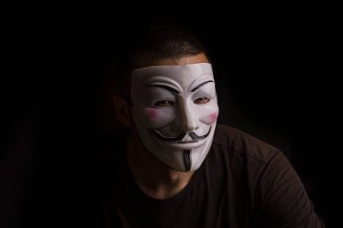 Karanlık bir odada kimliği belirsiz bir adam Guy Fawkes maskesi takıyor..
