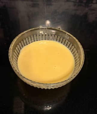 Koyu bir mutfak yüzeyinde şeffaf cam bir kâse içinde sıcak sarı krem. Yumuşak yansımaları ve pürüzsüz dokusu olan en az besin bileşimi.