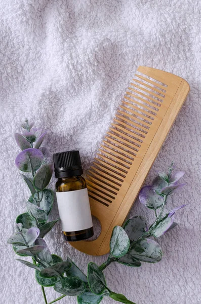 Top view of bottle of herbal oil for hair, green leaves and wooden comb on the white towel