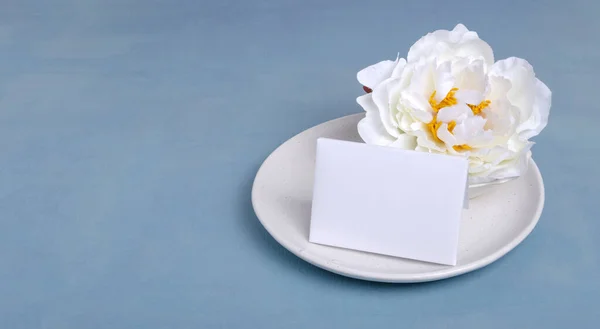 Closeup of white blank paper card, white peony on the plate, blue empty table
