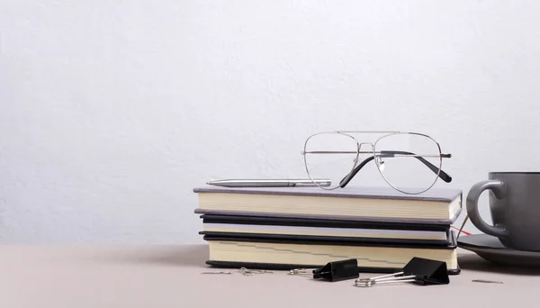 Closeup of stack of notebooks, eye glasses, cup of coffee, stationary binder clips on the desk against white empty wall