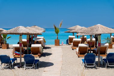 Nydri - İyon Adaları - Yunanistan - 08-09-2025: Ahşap yol, saman şemsiyeler ve şezlonglarla kumlu sahil boyunca uzanır, berrak gökyüzü altında turkuaz denize bakan, su altında şişme oyuncakla.