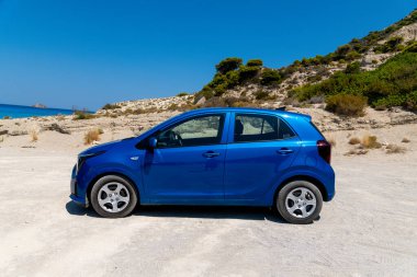 Nydri - İyon Adaları - Yunanistan - 08-09-2025: Açık gökyüzü ve parlak güneş ışığı altında seyrek bitki örtüsü bulunan kayalık bir sahil yamacının yakınındaki kumlu zeminde park edilmiş mavi bir kompakt araba.