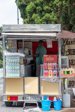 Rom - Lazio - İtalya - 08-21-2025: Bir yol kenarında seyyar atıştırmalık büfesi, içecekler ve paketlenmiş yiyeceklerle dolu bir şekilde açık dururken, bir kişi ağaçların altında ve bir lambanın altında dinleniyor.