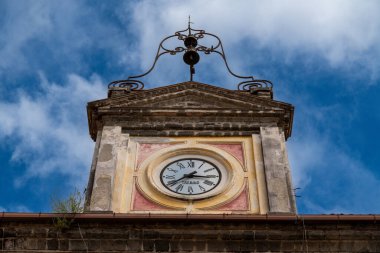 Sessa Aurunca - Campania - İtalya - 08-29-2025: Yıpranmış taş saat kulesi ön cephesi yuvarlak ve süslü metal çan yapısı dağınık bulutlu parlak mavi gökyüzüne doğru yükseliyor.