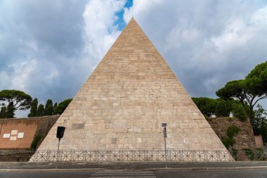 Önden Cestius-Piramide gösteriliyor, büyük taş blokları dik üçgen bir yüz oluşturuyor, anıtın etrafında ağaçlar ve bulutlar var..
