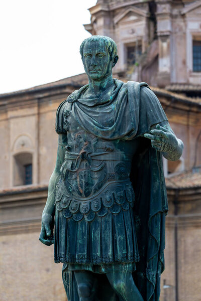 A close view of a bronze statue of Julius Caesar in armor shows raised detail on the cuirass and mantle, the figure poses frontally with blurred architecture.