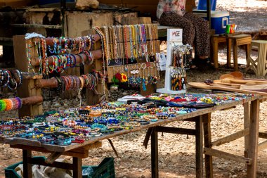 Taraf - Antalya - Türkiye - 07-07-2023: Renkli boncuk bilezikler ve kolyeler, anahtarlıklar ve oyulmuş kaşıkların yanı sıra açık bir tezgahtaki bir masayı, arkasında oturan bir kişiyle kaplıyor.