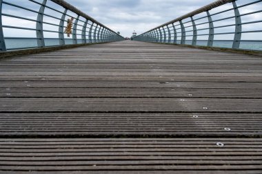 Groemitz - Holstein - Almanya - 06-28-2025: Denizin üzerinde eğri büğrü metal korkulukları olan geniş bir ahşap iskelenin alçak açılı görüntüsü, uzaktaki insanlara doğru gidiyor ve önümüzde küçük bir kule var.