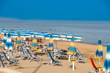 Foce Varano - Apulia - İtalya - 08-28-2025: Mavi ve beyaz plaj şemsiyeleri sakin bir denizin yanındaki kumlu sahilde güneşlenirler, açık gökyüzü altında sisli bir kıyı şeridi vardır..