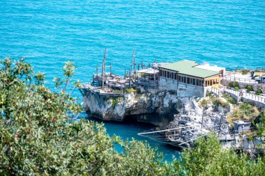 Vieste - Apulia - İtalya - 08-29-2025: Ahşap balıkçılık platformları ve küçük bir bina aşağıdaki turkuaz suyun üzerindeki kayalık bir uçurumun üzerinde, ön planda bulanık ağaçlar var..