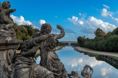 Reggia di Caserta - Campania - İtalya - 08-30-2025: Barok taş heykelleri, Caserta Kraliyet Sarayı 'na uzanan mavi bulutların altındaki ağaçlık yollar arasındaki uzun bir kanala tepeden bakıyor.