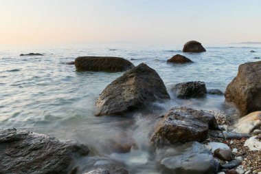 Önünde siyah kayalarla denizde gün batımı. Uzun kepenk açma zamanı. Kızıl gökyüzü ile bulanık deniz