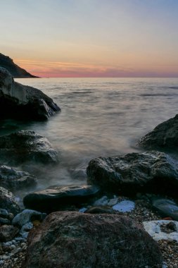 Önünde siyah kayalarla denizde gün batımı. Uzun kepenk açma zamanı. Kızıl gökyüzü ile bulanık deniz