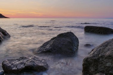 Önünde siyah kayalarla denizde gün batımı. Uzun kepenk açma zamanı. Kızıl gökyüzü ile bulanık deniz