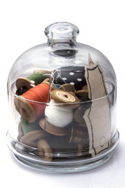 colored thread for sewing on wooden spools, needles, buttons, metal thimble stacked in transparent glass jar, sewing, hobby on white background