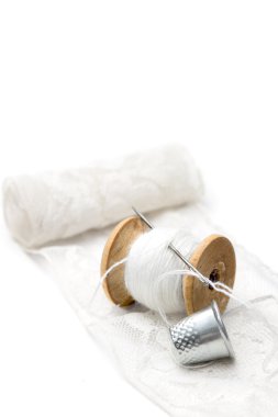 roll of white lace, a wooden spool of white cotton thread for sewing with needle and a metal thimble on white background