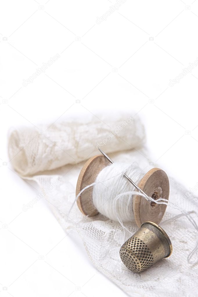 roll of white lace, a wooden spool of white cotton thread for sewing with needle and a metal thimble on white background