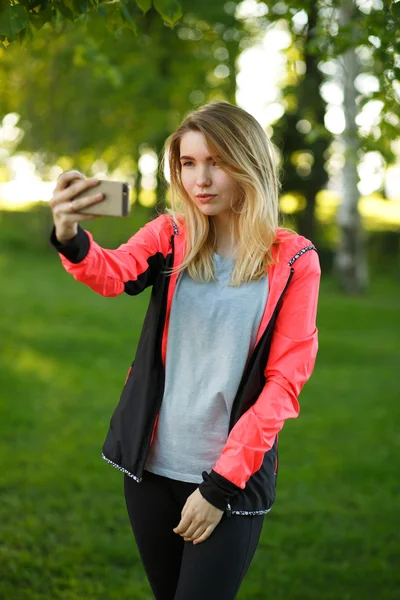 Çekici kadın atlet selfie yeşil park güneşli yaz günü yapıyor. O ayakta ve gülümseyerek. Kadın başparmak vazgeçmeden ve mutlu bir şekilde cep telefonu arıyorum