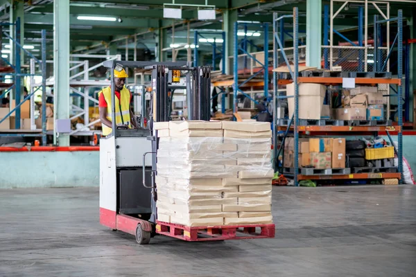 African American factory man or warehouse worker stand on forklift during work in workplace area to transfer pack of product. Concept of good management work in delivery industrial business.