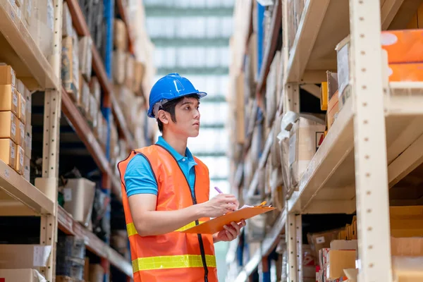 Warehouse man or factory worker with blue hard hat and uniform stand between shelves and check package in workplace. Concept of good management and happiness of staff during work industrial business.