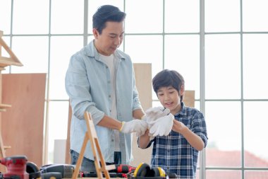 Asyalı baba, evinde ahşap işçiliğiyle çalışmadan önce oğluna eldiven giymesinde yardım ediyor. Hobi ile iyi ilişki kavramı ya da mutlu aile aktiviteleri.