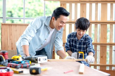 Asian father look carefully son use saw with timber in their workplace of carpentering with happy emotion. Asian family concept to stay at home and enjoy good relationship hobby together. clipart
