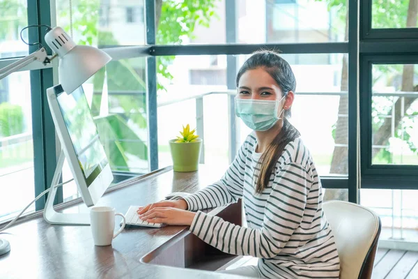 Asian woman with hygiene mask sit in working room in front of computer and look at camera during work at home with day light.