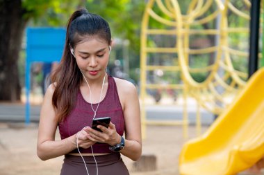 Asyalı kadın portresi, parkta ya da bahçede sabah güneşli egzersiz yaparken cep telefonu kullanmaya ve oyun parkının yakınında kalmaya konsantre olur..