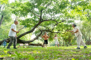 Asyalı üst düzey erkek ve kadın halk parkında mutlu mesut badminton oynarlar ve iki küçük kız neşelenir..
