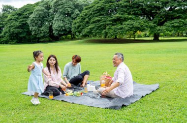 Asyalı küçük kız annesinin, babasının ve dedesinin etrafında koşuyor. Gün ışığı ve mutlulukla halka açık parkta birlikte piknik yapıyorlar..