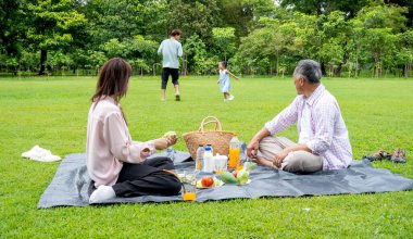 Anne ve büyükbaba halk parkında çimenlerde oturur ve baba ve torunun gün ışığıyla birlikte oynamaktan zevk aldıklarına bakarlar..