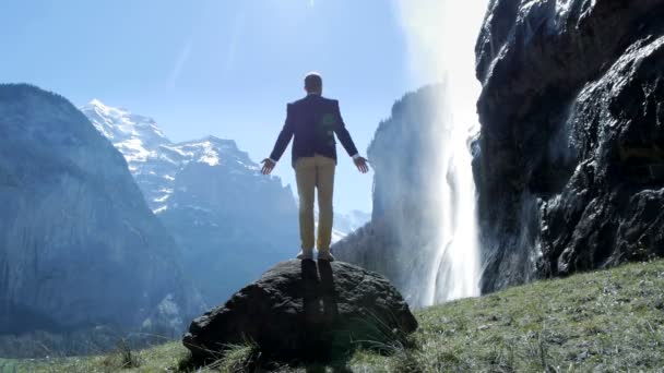Человек стоит на камне и смотрит на водопад . — стоковое видео