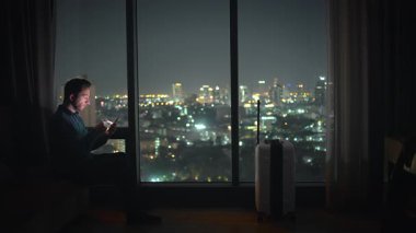 Düşünceli bir adam otel odasında oturur ve gece vakti aydınlatan çarpıcı şehir manzarasını yansıtır.
