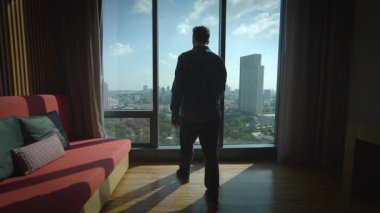 Şık, modern bir odadan geniş şehir manzarasına dikkatle bakan bir adamın çarpıcı silüeti.