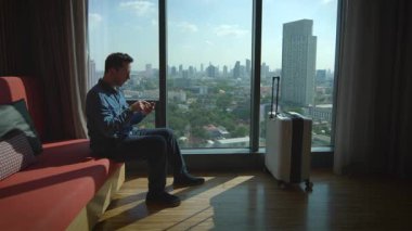 A man sits by the window, enjoying the stunning city skyline view while focused on his mobile device