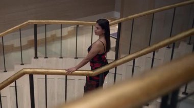 woman standing on stair landing, hand on brass railing, looking across lobby, formal dress,