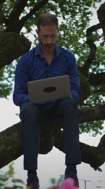 Bir adam açık havada dizüstü bilgisayar kullanarak sağlam bir ağaç dalına rahatça oturmuş yaratıcılık ve yenilik sergiliyor.