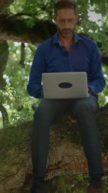 Bir adam, sakin bir doğal ortamda, dayanıklı bir ağaç dalında otururken dizüstü bilgisayarı üzerinde rahatça çalışır.
