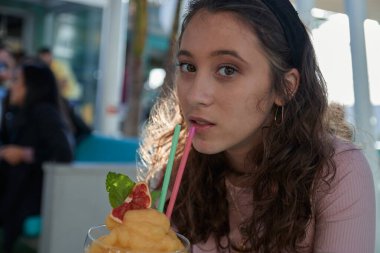 Güneşli bir günde barda mango kokteyli içen bir kız pembe bir tişört giyiyor.