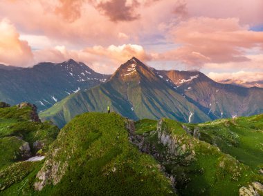 Sırt çantalı bir yürüyüşçü, yeşil bir dağın zirvesinde, günbatımı gökyüzünün arka planına karşı. Başarılı yükseliş
