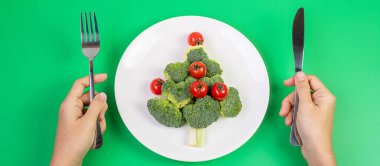 Beyaz tabakta organik sebzelerden oluşan Noel ağacı; Brokoli ve yeşil arka planda çatal ve bıçaklı domatesler. Mutlu Noeller ve mutlu yeni yıl partisi, yeni sen, sağlıklı yemek ve vejetaryen konsepti