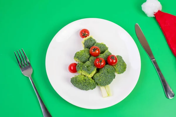 Christmas tree of organic vegetables on white plate; Broccolis and tomatoes with fork and knife on green background. Merry Xmas and happy New Year party, New You, Healthy food and vegetarian concept