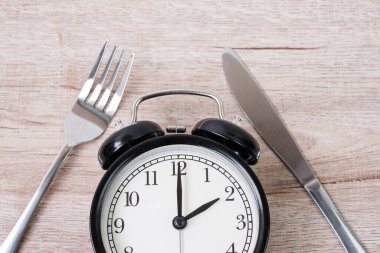 Ahşap masa zemininde çatal ve bıçak bulunan üst görüşlü çalar saat. Aralıklı oruç, Ketogenik diyet, kilo kaybı, yemek planı ve sağlıklı gıda konsepti